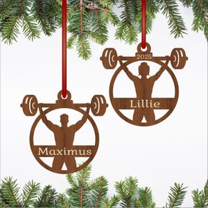 May include: Two wooden Christmas ornaments with a weightlifting theme. Each ornament features a silhouette of a person lifting a barbell inside a circle. One ornament says "Maximus", the other says "Lillie" and "2025". Both are suspended by red ribbons.