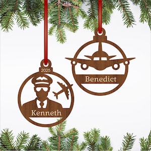 May include: Two wooden Christmas ornaments suspended by red ribbons. One features a pilot silhouette with the name "Kenneth" and the year "2025". The other displays an airplane design with the name "Benedict". Both are set against a backdrop of evergreen branches.