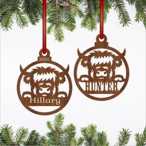 May include: Two wooden Christmas ornaments with a Highland cow design. One ornament has the name "Hillary" and the year "2025". The other ornament has the name "Hunter". Both are suspended by red ribbons against a backdrop of evergreen branches.