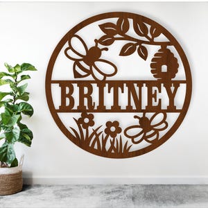 Può includere: Un cartello circolare in legno marrone con il nome "BRITNEY" ritagliato a grandi lettere. Il cartello presenta ritagli di api e fiori, insieme a un alveare e fogliame. Il cartello è montato su un muro bianco.