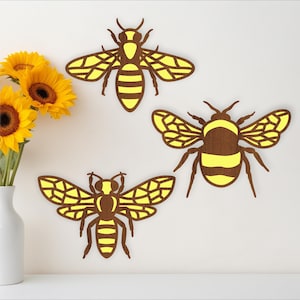 May include: Three wooden bee cutouts with yellow and brown details are displayed on a white wall. A bouquet of sunflowers in a white vase sits on a white surface. The bees have intricate wing patterns.
