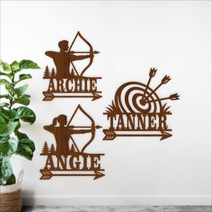 May include: Wooden wall art featuring archers and a target. The archers are depicted with bows and arrows, with the names "Archie" and "Angie" below them. A target with the name "Tanner" is also included. The art is in a brown color.