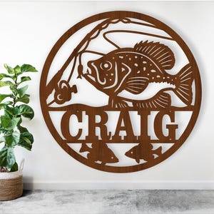 May include: A circular, brown metal wall art piece featuring a fish caught on a fishing line. The name "CRAIG" is cut out below the fish, with two smaller fish silhouettes at the bottom. The art is mounted on a white wall.