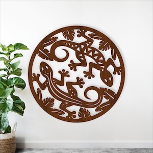 May include: A circular, brown wooden wall art piece featuring two stylized gecko lizards and leaf accents. The lizards are intricately cut out, creating a decorative and artistic design. The artwork is mounted on a white wall.