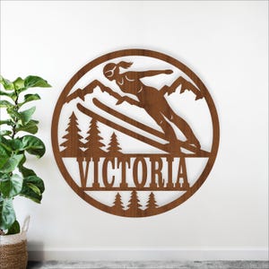 May include: A round, wooden wall art piece featuring a skier in action, mountains, and pine trees. The name "VICTORIA" is displayed in large letters. The artwork is a warm brown color and is designed for wall decor.