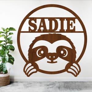 以下が含まれることがあります： サディーという名前が書かれた円の中に、茶色の木製のナマケモノの切り抜きがあります。ナマケモノはかわいい表情をして、円から顔をのぞかせています。