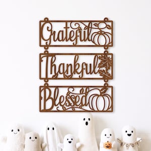May include: A brown laser-cut wooden wall sign consisting of three rectangular panels connected vertically. The panels feature the words Grateful, Thankful, and Blessed in a cursive font, decorated with pumpkin and autumn leaf cutouts.