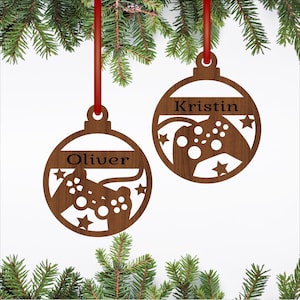 May include: Two wooden Christmas ornaments with red ribbons, each featuring a name and a video game controller design. One ornament says "Oliver," the other says "Kristin." The ornaments are brown and surrounded by star cutouts.