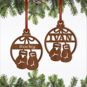 May include: Two wooden Christmas ornaments with a boxing theme. One ornament says "Rocky" and "2025", the other says "IVAN". Each ornament features a pair of boxing gloves. The ornaments are suspended by red ribbons and surrounded by evergreen branches.