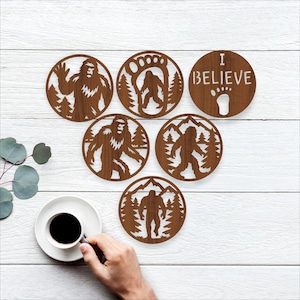 May include: Six brown wooden coasters with Bigfoot designs. Each coaster features a different Bigfoot illustration, including a footprint and the words "I BELIEVE". The coasters are round and have a rustic, natural look, perfect for a cabin or nature-themed decor.