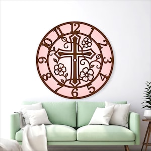 May include: A decorative wall clock featuring a cross design with floral accents. The clock has a brown frame and a pink background. The numbers are visible around the clock face. The clock is mounted on a white wall above a green sofa.