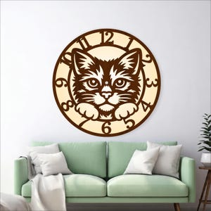 May include: A wooden cat-themed wall clock. The clock features a detailed cutout of a cat's face at the center, surrounded by a circular frame with numbers. The cat's face is brown, and the background is a lighter beige color. The clock is hanging on a white wall.