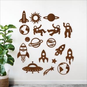 Puede incluir: Juego de decoración de pared de madera con temática espacial. Incluye un cohete, planetas, astronautas, el sol y una estrella fugaz. Los recortes marrones están dispuestos en una pared blanca, creando un tema lúdico y educativo.