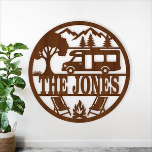 Pode incluir: Uma placa redonda de metal castanho com tema de acampamento. O design inclui uma autocaravana, montanhas, árvores, uma fogueira e duas espreguiçadeiras. O texto "THE JONES" é exibido no centro.