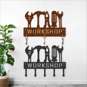 May include: Two workshop-themed key holders. The top one is brown, and the bottom one is black. Each features cut-out tool silhouettes above the word "WORKSHOP" and has key hooks below. The brown key holder has three keys hanging from it.