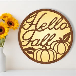 May include: A round wooden sign with the words "Hello Fall" in a decorative font. The sign is illuminated from behind, creating a warm glow. There are two stylized pumpkins with leaves on the bottom of the sign.