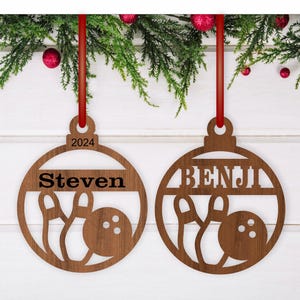 May include: Two wooden Christmas ornaments shaped like bowling balls with the names "Steven" and "Benji" on them. The ornaments are brown and have a white background.
