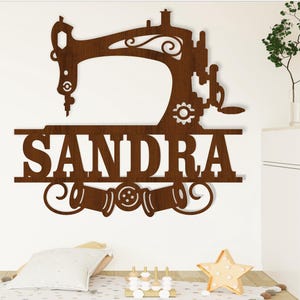 May include: Brown wooden wall decor featuring a silhouette of a sewing machine with the name "SANDRA" underneath. The sewing machine has a decorative scroll design and there are three spools of thread below the name.