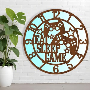 May include: A brown wooden clock with a blue background. The clock face features a design of two video game controllers, stars, and the text "Eat Sleep Game".