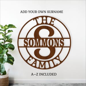 May include: A brown metal family name sign with the text "THE SOMMONS FAMILY" and a large letter "S" in the center. The sign is circular with the text "ADD YOUR OWN SURNAME" and "A-Z INCLUDED" above and below.