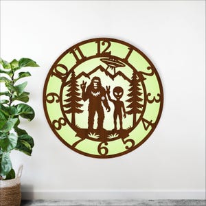 May include: A round wooden clock with a brown frame and a light green center. The clock features a Bigfoot and alien design with a UFO above a mountain range. The numbers are in a brown, stylized font.