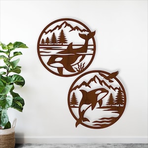May include: Two circular wooden wall art pieces featuring orcas, mountains, and trees. One shows an orca swimming, the other shows an orca jumping. The art is a warm brown color and is mounted on a white wall.