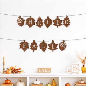 May include: A decorative banner consisting of two strings of brown wooden cutouts shaped like autumn leaves and acorns. The top string spells HELLO and the bottom string spells FALL, with each letter cut out from the centre of the wooden shapes.