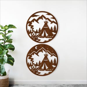 May include: Two circular wooden wall art pieces. Each features a mountain range, a teepee, a campfire, and trees. The top piece has a sun, and the bottom piece has a crescent moon. The art is a warm brown color.
