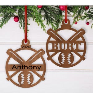 Op de afbeelding: Twee houten kerstversieringen in de vorm van honkballen met gekruiste knuppels. De versieringen zijn bruin en hebben een witte honkbal met rode stiksels in het midden. De bovenste versiering heeft de naam "BURT" bovenaan geschreven en de onderste versiering heeft de naam "Anthony" bovenaan geschreven. Het jaar "2024" staat op de bovenste versiering.