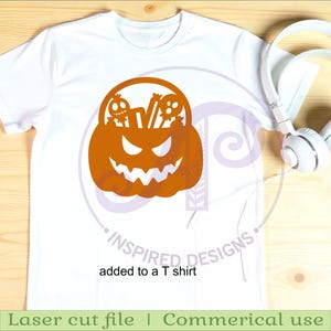 Puede incluir: Camiseta blanca con un dise&ntilde;o de calabaza naranja con tem&aacute;tica de Halloween, con una cara tallada y dos peque&ntilde;as figuras de calaveras. El texto "added to a T shirt" est&aacute; debajo del dise&ntilde;o. Auriculares blancos a la derecha.