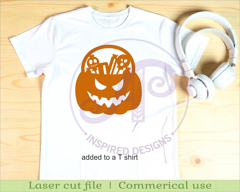Puede incluir: Camiseta blanca con un dise&ntilde;o de calabaza naranja con tem&aacute;tica de Halloween, con una cara tallada y dos peque&ntilde;as figuras de calaveras. El texto "added to a T shirt" est&aacute; debajo del dise&ntilde;o. Auriculares blancos a la derecha.