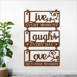 May include: A brown metal wall art piece with three rectangular plaques. The top plaque reads "Live Every Moment", the middle plaque reads "Laugh Every Day", and the bottom plaque reads "Love Beyond Words". Decorative floral and heart accents.
