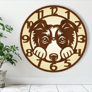 Op de afbeelding: Een houten klok met een bruine rand en een crèmekleurige wijzerplaat. De klok heeft een zwart-witte border collie hondenkop met zijn poten die de cijfers 10 en 4 bedekken. De cijfers 1 tot en met 12 staan op de wijzerplaat van de klok.