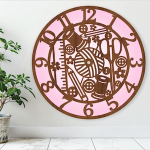 Puede incluir: Un reloj de madera rosa y marrón con temática de costura. La esfera del reloj presenta varias herramientas de costura, incluyendo tijeras, hilo, agujas y botones. Los números en la esfera del reloj están en una fuente decorativa.