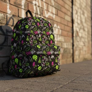 May include: Black backpack with a repeating pattern of neon green and pink doodles, including skulls, smiley faces, and text. The backpack has a top handle and two shoulder straps. It is leaning against a brick wall.