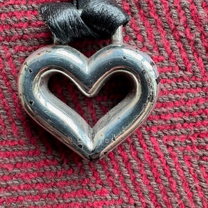 Puede incluir: Un colgante de plata en forma de coraz&oacute;n con un cord&oacute;n de cuero negro. El colgante tiene una forma de coraz&oacute;n recortada en el centro.