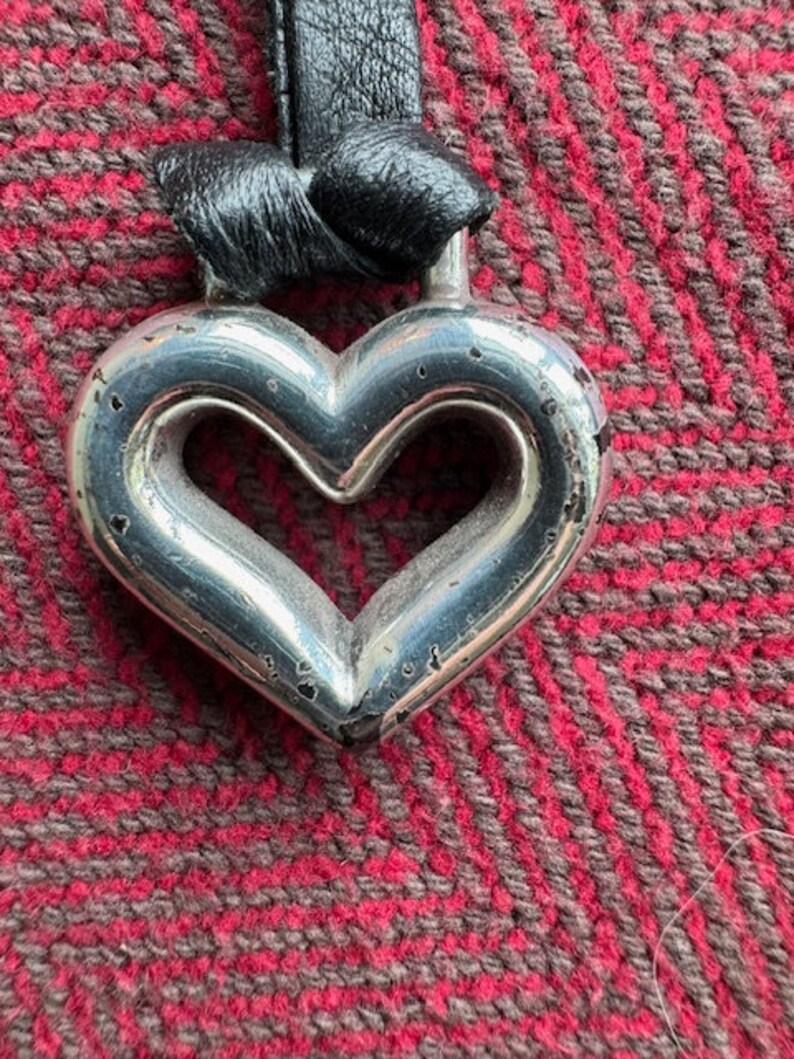 Puede incluir: Un colgante de plata en forma de coraz&oacute;n con un cord&oacute;n de cuero negro. El colgante tiene una forma de coraz&oacute;n recortada en el centro.
