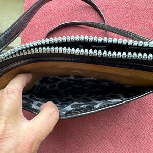 Puede incluir: Un bolso de hombro de cuero negro con cierre de cremallera y forro de estampado de leopardo. El bolso tiene una sola correa.