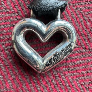 Puede incluir: Un colgante de plata en forma de coraz&oacute;n con la palabra "Brighton" grabada. El colgante est&aacute; unido a un cord&oacute;n de cuero negro.