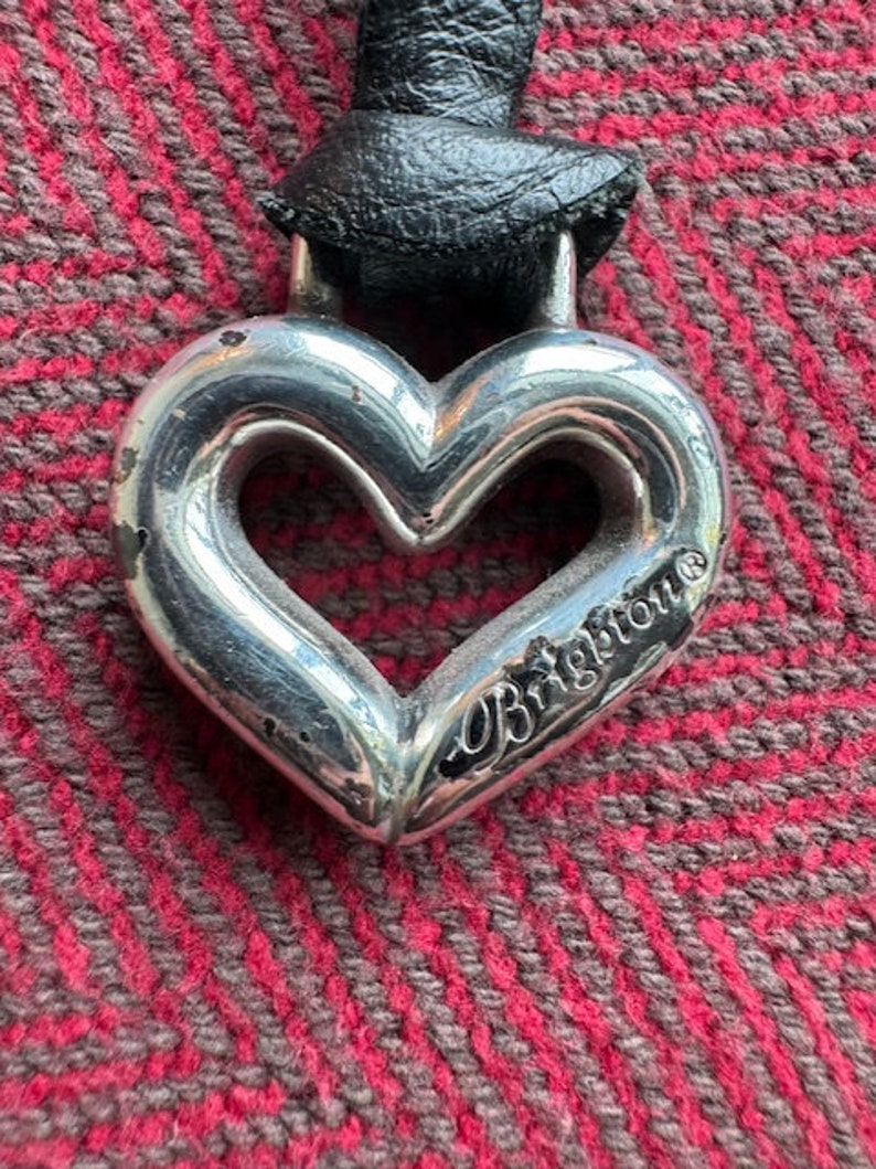 Puede incluir: Un colgante de plata en forma de coraz&oacute;n con la palabra "Brighton" grabada. El colgante est&aacute; unido a un cord&oacute;n de cuero negro.