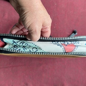 Puede incluir: Una bolsa blanca con cremallera con un estampado colorido que incluye el texto "Taste the Trip" y un coraz&oacute;n rojo. La bolsa tiene una cremallera negra y un forro marr&oacute;n.