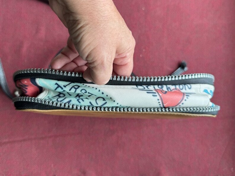 Puede incluir: Una bolsa blanca con cremallera con un estampado colorido que incluye el texto "Taste the Trip" y un coraz&oacute;n rojo. La bolsa tiene una cremallera negra y un forro marr&oacute;n.