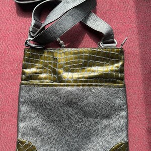 Op de afbeelding: Een grijze leren schoudertas met een groene krokodil-ge&euml;mbosseerde klep en accenten aan de onderkant. De tas heeft een lange verstelbare schouderband.