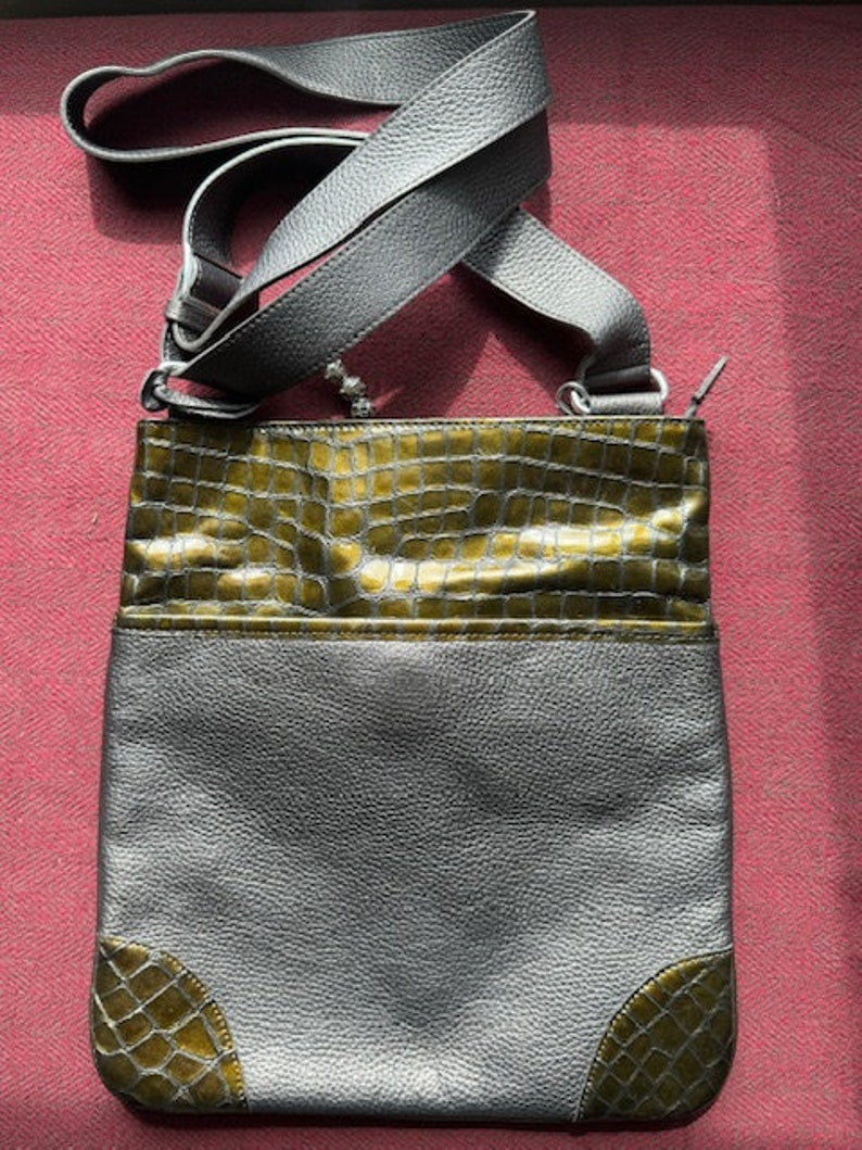 Op de afbeelding: Een grijze leren schoudertas met een groene krokodil-ge&euml;mbosseerde klep en accenten aan de onderkant. De tas heeft een lange verstelbare schouderband.