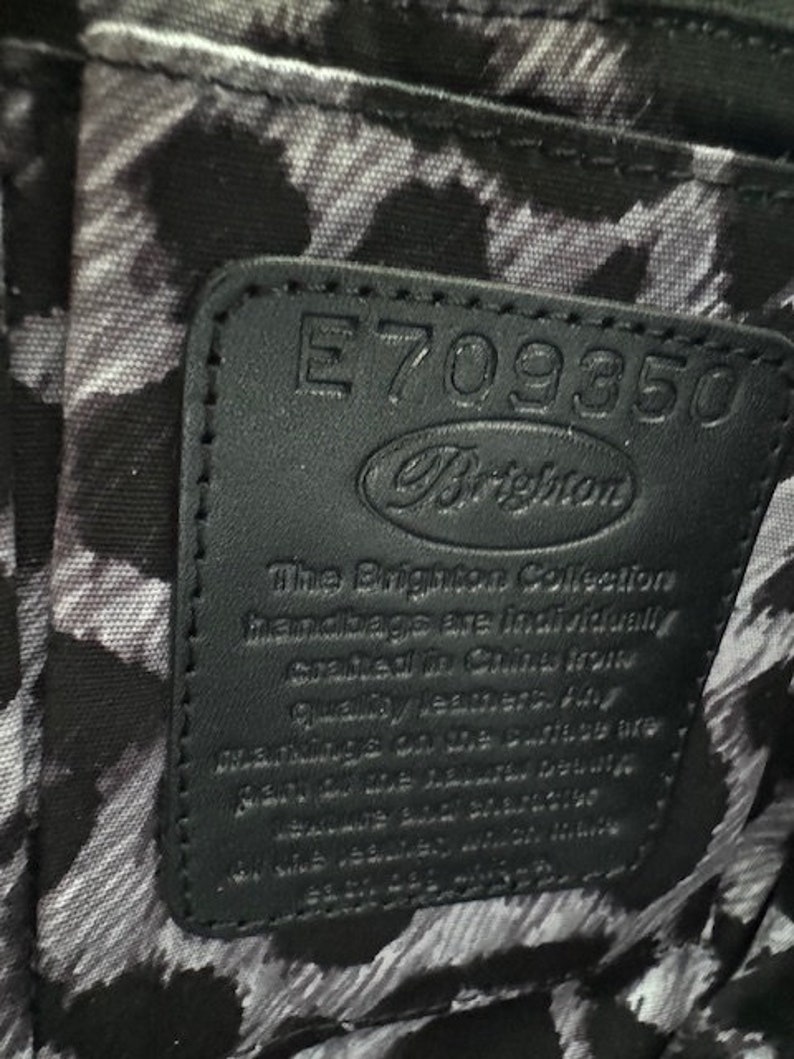 Puede incluir: Etiqueta de cuero negro con costuras blancas y el texto "E709350 Brighton The Brighton Collection handbags are individually crafted in China from quality leathers. Any marks on the surface are made by the leather itself and should not be seen as a defect. Made in China."