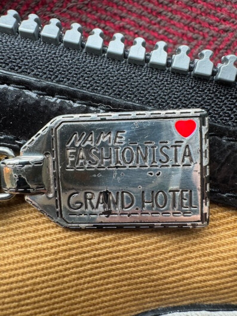 Puede incluir: Una etiqueta de metal plateado con el texto "NAME FASHIONISTA GRAND HOTEL" y un coraz&oacute;n rojo. La etiqueta est&aacute; unida a una cremallera negra.