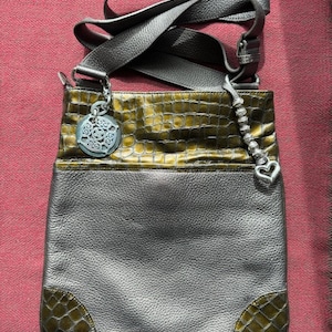 Op de afbeelding: Een grijze leren schoudertas met een groen krokodilpatroon. De tas heeft een zilveren hanger met een decoratief ontwerp.