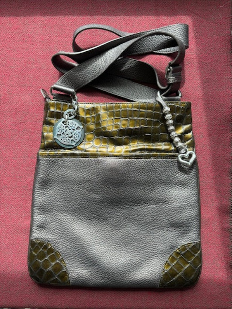 Op de afbeelding: Een grijze leren schoudertas met een groen krokodilpatroon. De tas heeft een zilveren hanger met een decoratief ontwerp.