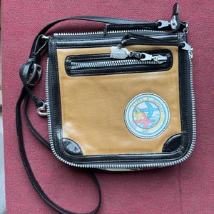 Puede incluir: Un bolso bandolera de cuero marr&oacute;n y negro con cierre de cremallera y un parche con el texto "Little & Souvenir" y un gr&aacute;fico de tres aviones.