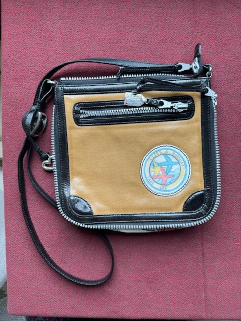 Puede incluir: Un bolso bandolera de cuero marr&oacute;n y negro con cierre de cremallera y un parche con el texto "Little & Souvenir" y un gr&aacute;fico de tres aviones.